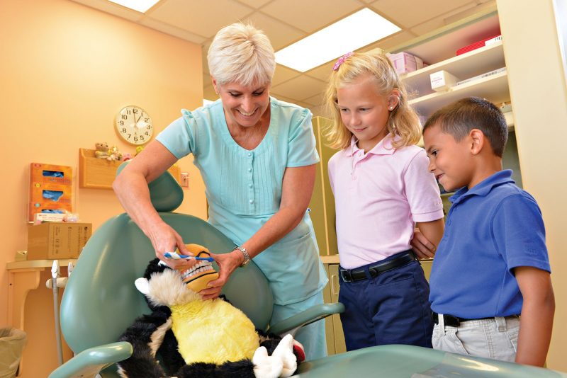 Female dentist using a toy to show two young Milton Hershey School students how to brush teeth.