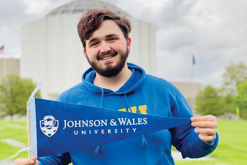 MHS student holding Johnson & Wales University banner in front of Founder's Hall building