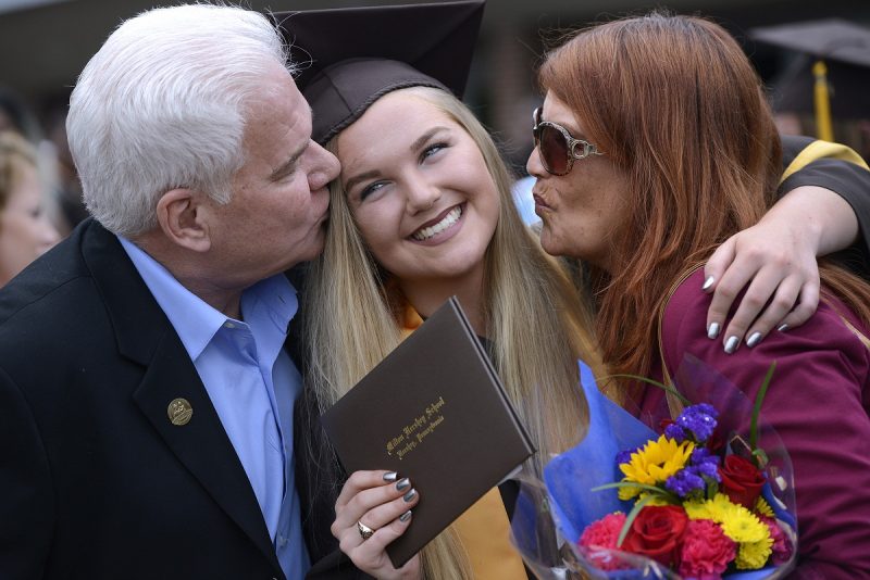 A father and mother each kiss a cheek of a female MHS student in cap and gown.